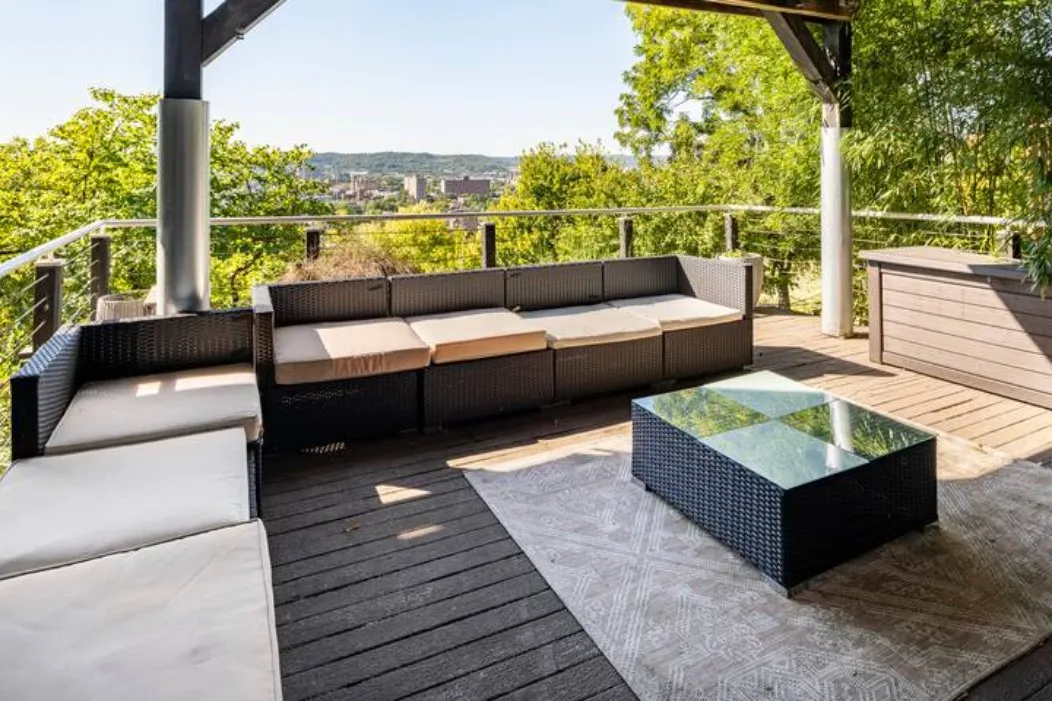 Covered deck with wicker sofas and a glass-topped table overlooking trees and the city